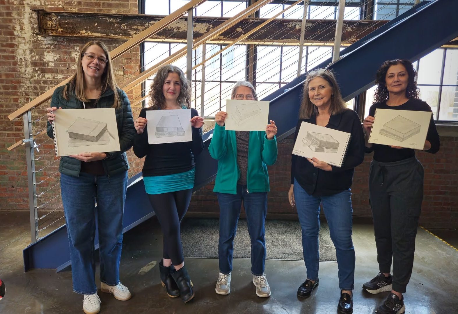 Five adult students standing in a line holding up sketchbooks with perspective drawings of books
