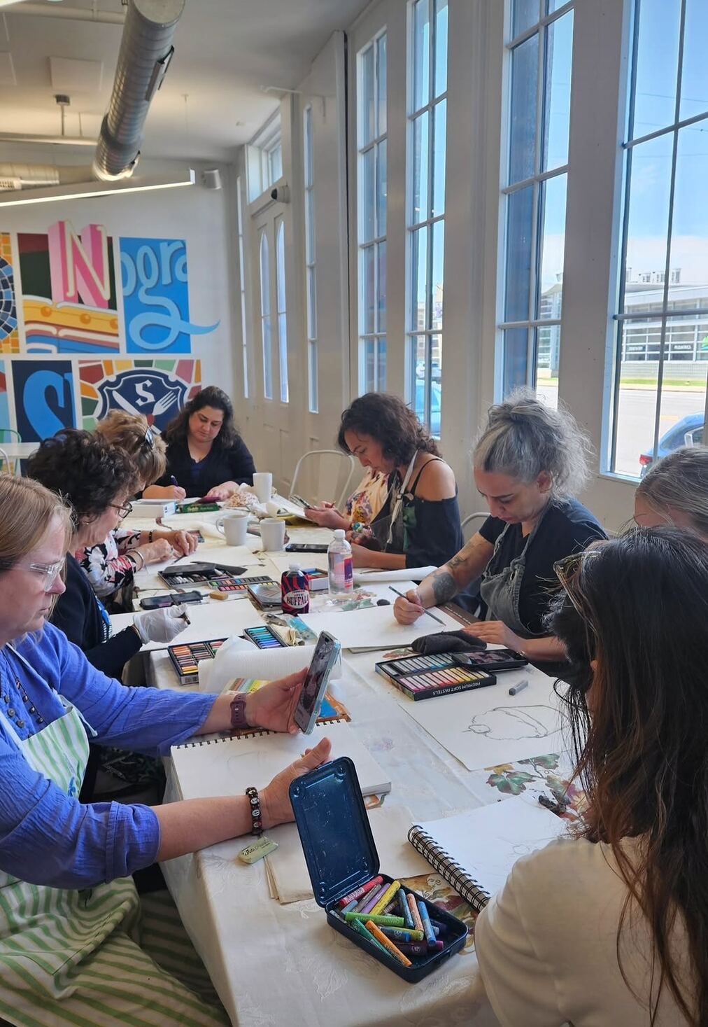 Adult students drawing with soft pastels at a bright studio table by tall windows