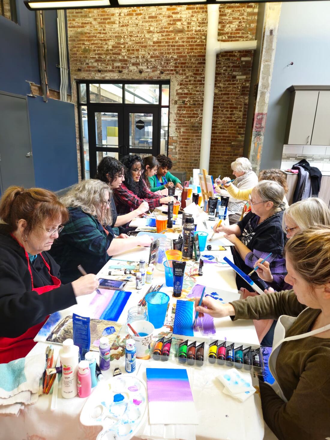 A group of adult students painting at a long table in a studio, canvases in early stages