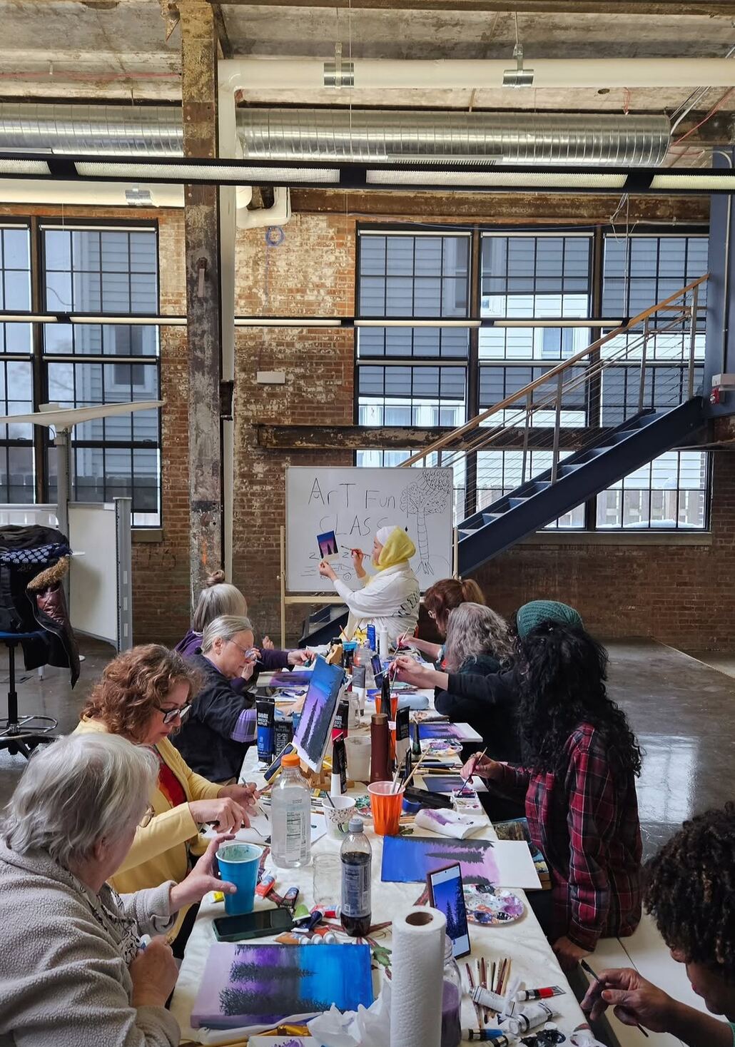 Tan demonstrating at a whiteboard while adult students paint at a long studio table
