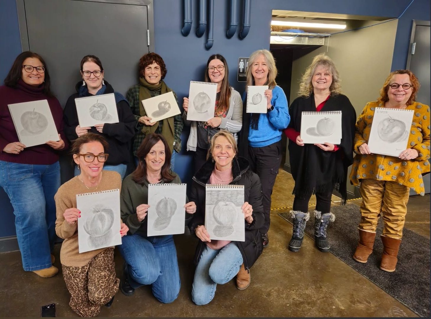 Ten adult students posing together holding up sketchbooks with shaded pencil drawings of apples